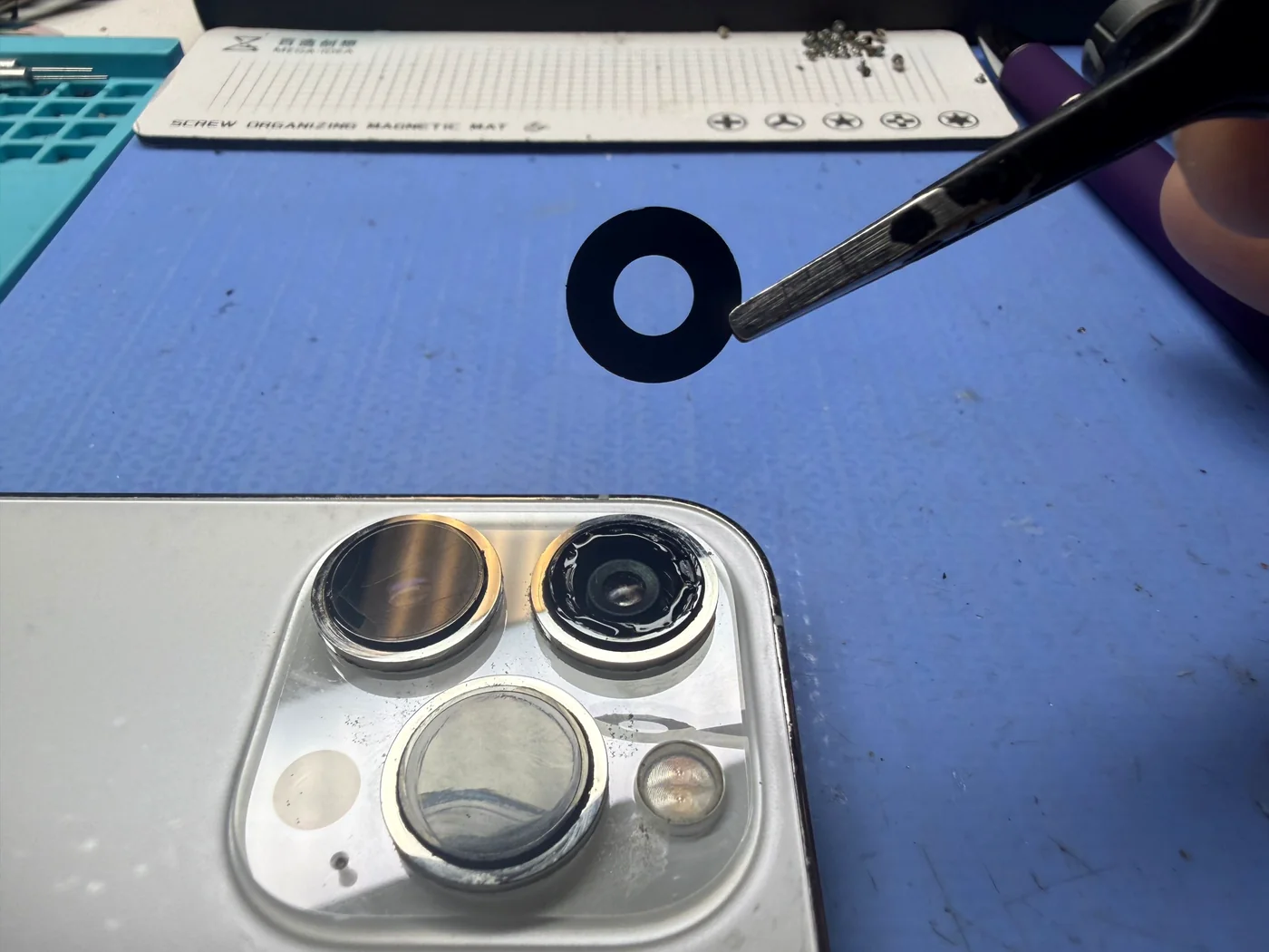 New genuine Apple camera lens cover glass held in tweezers above an iPhone 14 Pro with the old glass removed